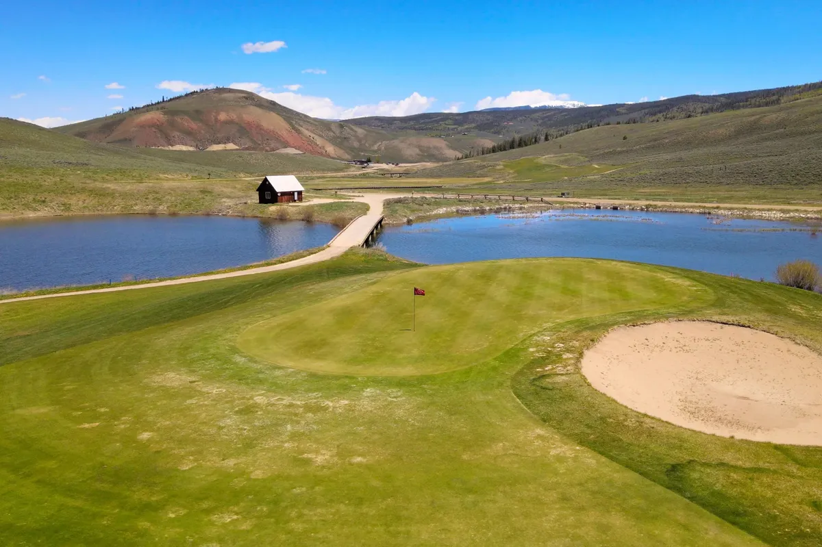 The hero image showcases a serene golf green bordered by a small wooden cabin, gentle water bodies, and a sand trap, all set against rolling hills that glow with the colors of sunset. The setting invites opportunities for golfing, relaxing picnics, and soaking in Colorado’s natural beauty.