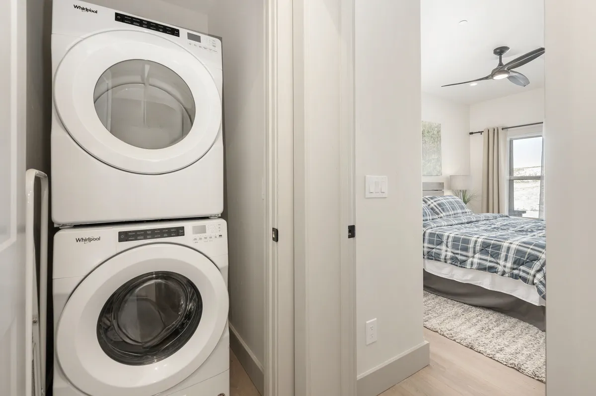The image shows a stacked washer and dryer next to a door leading into a bedroom, illustrating a compact, functional laundry area within the home. The scene is practical yet inviting, reinforcing the convenience for longer stays and active families.