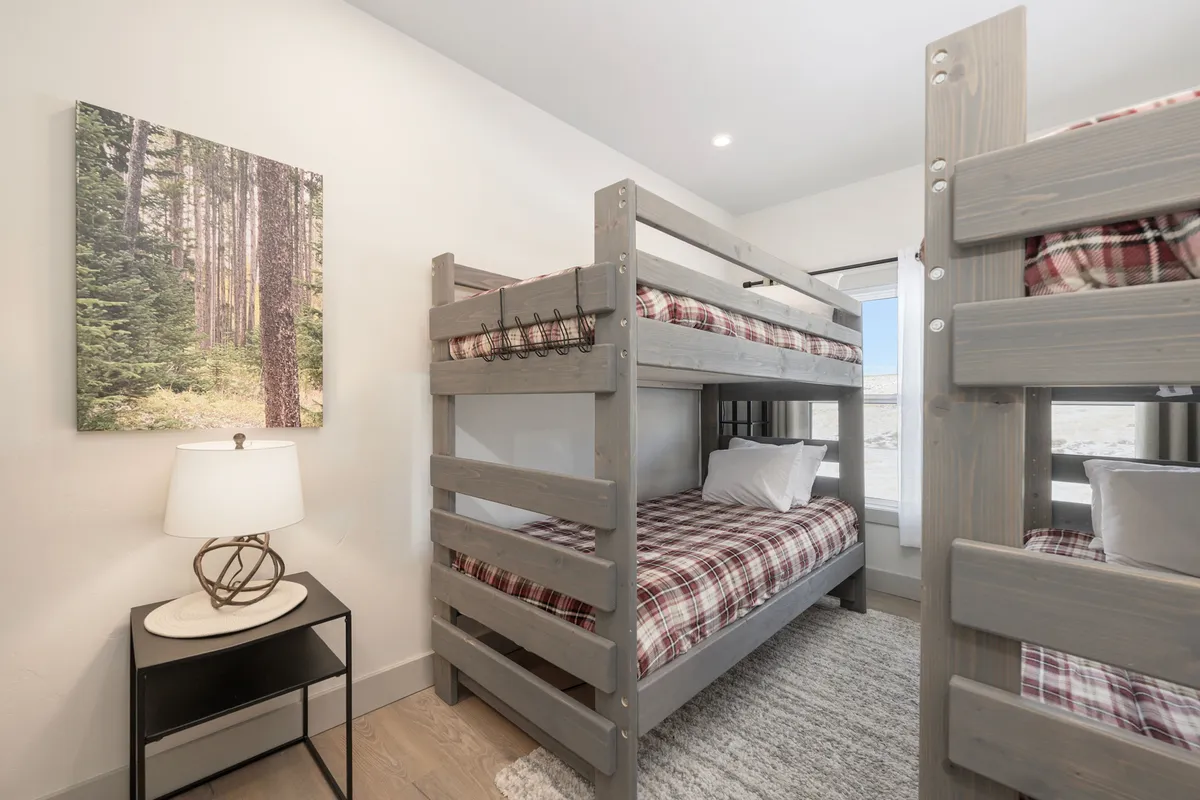 A playful, inviting bunk room features neatly made twin bunks, a landscape artwork above, and a side table with a lamp perfect for bedtime stories or reading. The setup is ideal for children or friends, evoking togetherness and fun.