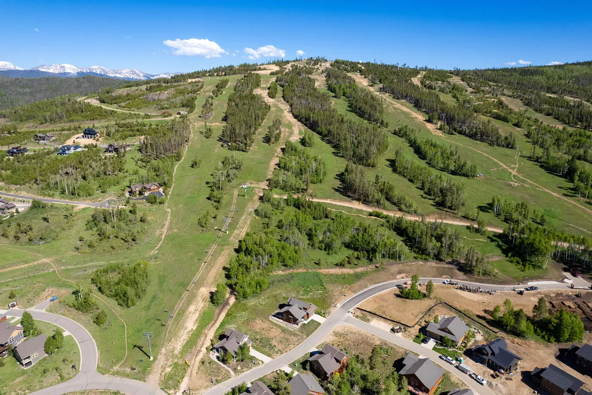 A panoramic view capturing ski slopes stretching across a majestic mountain range, with residential homes nestled at the base and a winding road leading into the adventure. The landscape hints at both winter and summer activities, with open meadows and towering peaks.