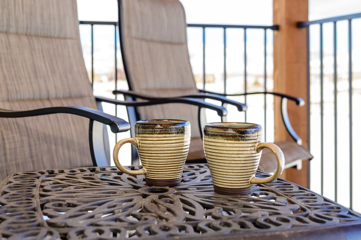 An inviting outdoor deck setup featuring two ceramic mugs on a decorative table, with outdoor chairs positioned to enjoy the surrounding scenery. The deck is bathed in soft natural light and offers a direct connection to the outdoors, suggesting a peaceful space to start the day or unwind in the evening.