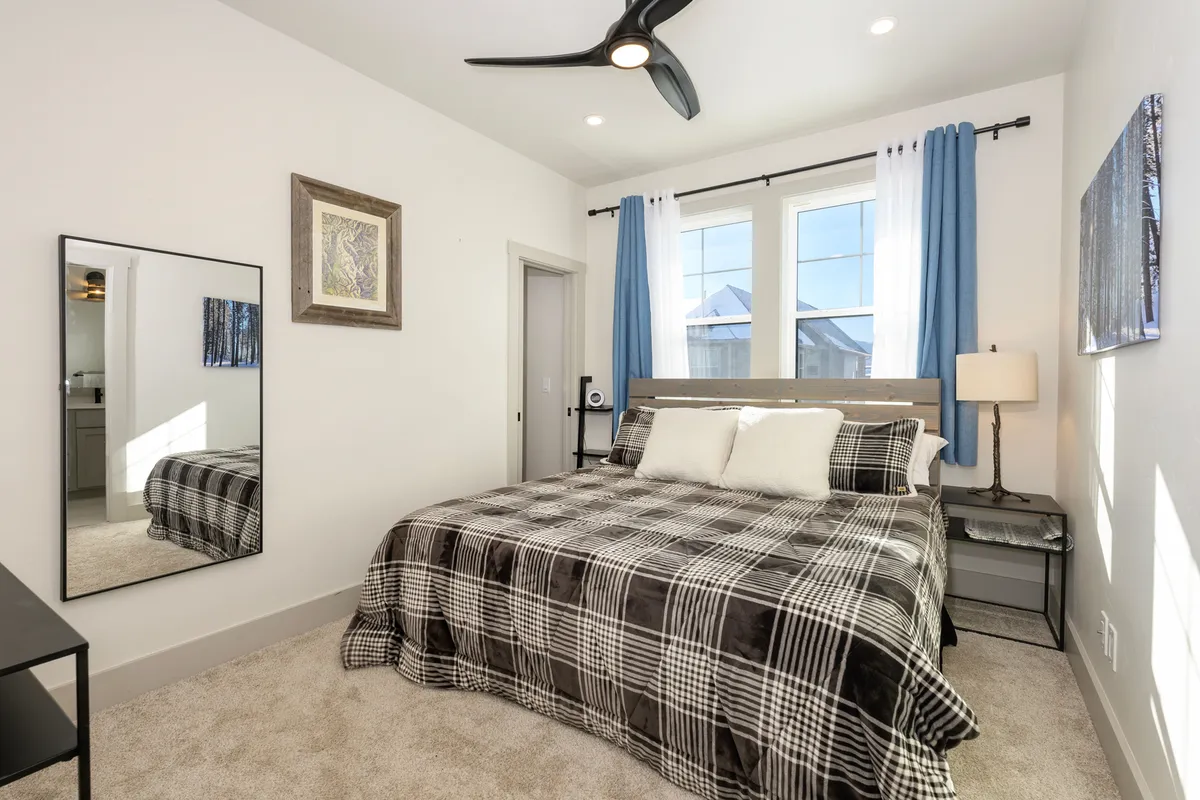 A king-sized bed with inviting plaid bedding is the centerpiece, flanked by a large mirror, blue curtains, and a nightstand with a warm lamp. Natural light filters gently into the room, creating a serene and restful atmosphere.