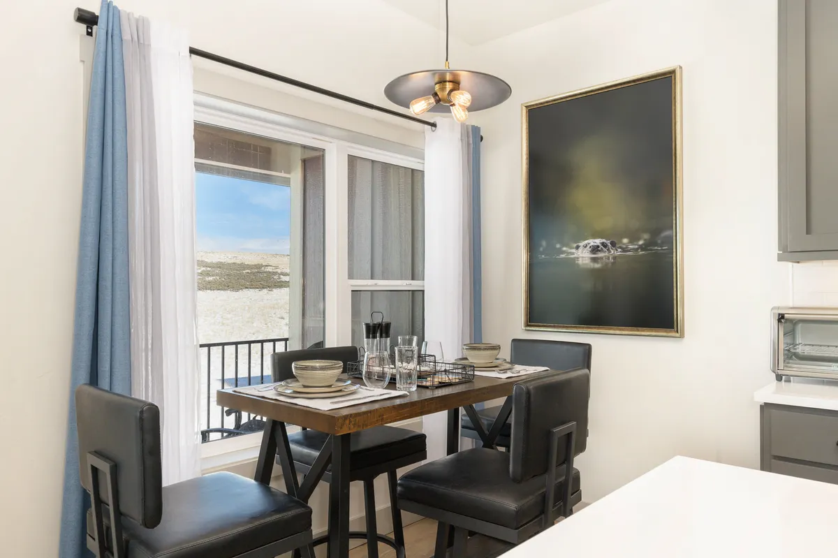 A bright, inviting dining area with a large window offering scenic views, a stylish wooden table set for a meal, and artwork on the walls. The space is designed for sharing meals and socializing, reflecting the comforts of home within a resort community.