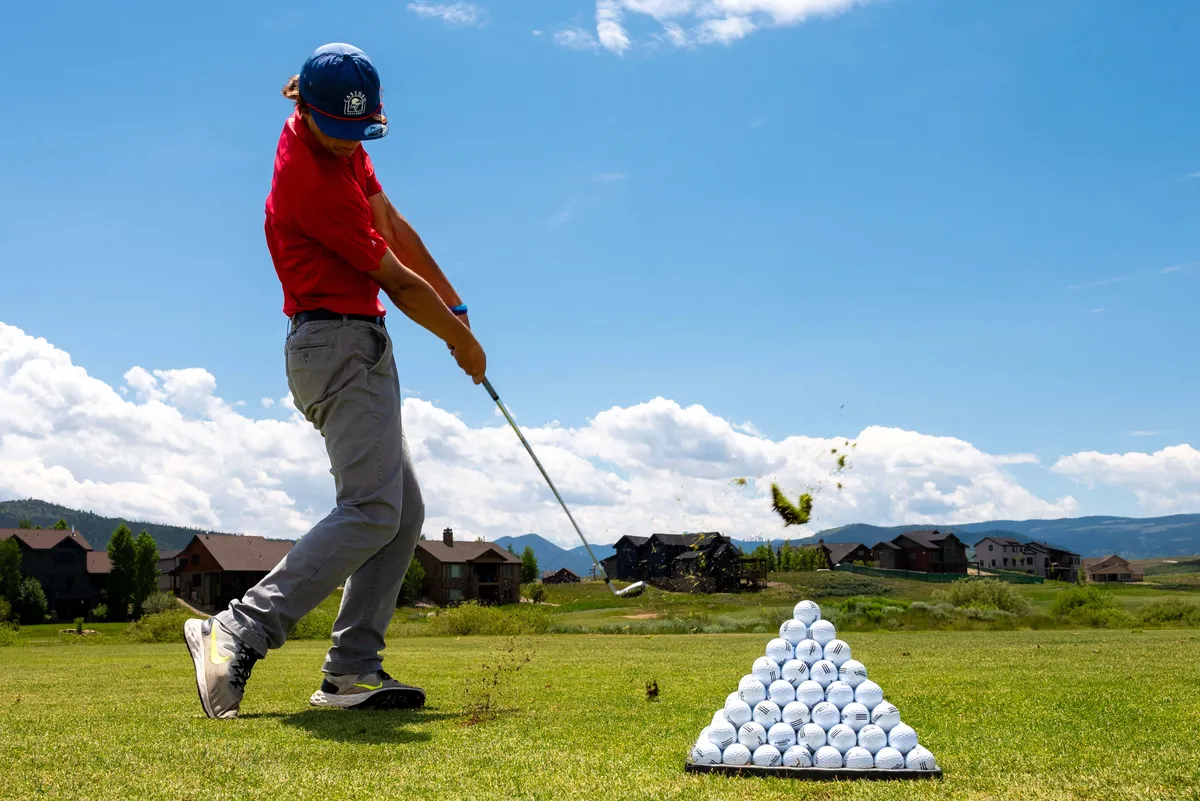 A golfer is in mid-swing on a practice area, with a pyramid of golf balls ready for use. The backdrop features stunning mountain vistas under a clear sky, emphasizing the connection between recreation and the natural landscape.